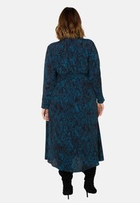 Långärmad klänning i golvlängd i mörk teal med ett ormsmönster. Har en figurnära midja och en flödande kjol. Bärs med svarta knähöga stövlar.