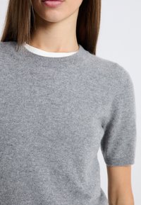 Szary sweter z krótkim rękawem wykonany z miękkiego dzianinowego materiału, z okrągłym dekoltem i subtelną fakturą, noszony na białym kołnierzyku koszuli.