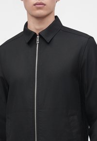 Homme portant une veste noire avec une fermeture éclair argentée sur le devant et un col pointu, visible du cou jusqu'au milieu du torse.