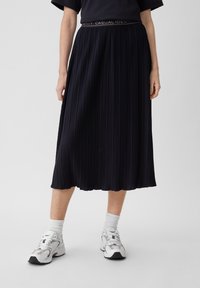 Gonna plissettata blu navy con finitura texturizzata e cintura elastica, caratterizzata da un logo bianco. Abbinata a calze sportive bianche e scarpe.