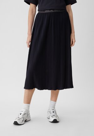 Gonna plissettata blu navy con finitura texturizzata e cintura elastica, caratterizzata da un logo bianco. Abbinata a calze sportive bianche e scarpe.