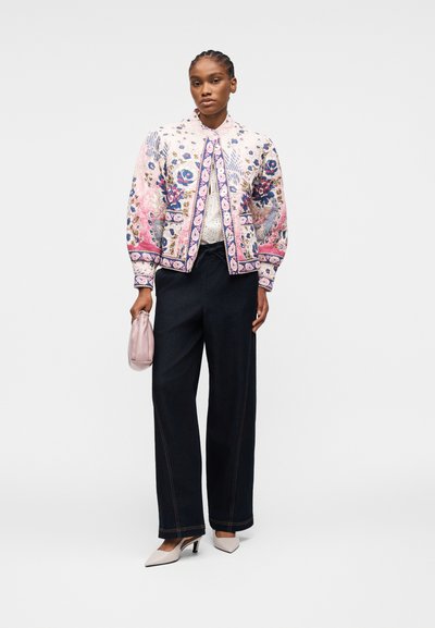 Femme portant une veste matelassée à motif floral, un haut blanc, un pantalon noir à jambes larges, des talons blancs, et tenant un petit sac à main rose.