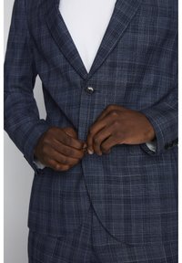 Marinblå rutigt kavaj med en enda knappstängning, texturerad tyg, och en vit skjorta under; händer som justerar kavajen.