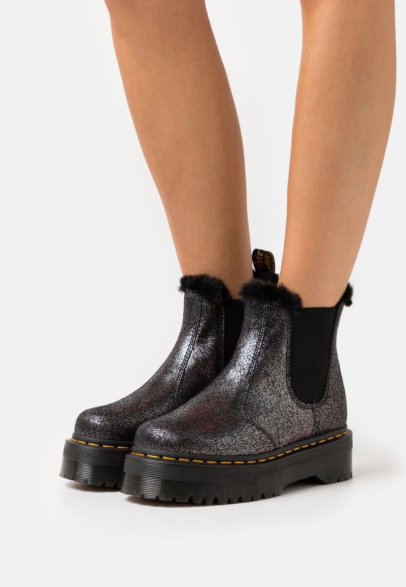 Black glitter ankle boots with faux fur trim, elastic side panels, chunky sole, and yellow stitching detail. Textured surface enhances visual appeal.