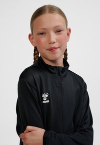 Svart sportjacka med dragkedja, tillverkad av slät tyg. Har en hög krage och en vit logotyp på vänster sida. Långa ärmar med åtsittande muddar.