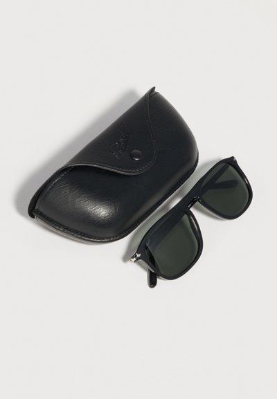 Μαύρα ορθογώνια γυαλιά ηλίου με σκοτεινούς φακούς δίπλα σε μαύρη θήκη από δέρμα με κουμπί από αστραπή σε λευκό φόντο.