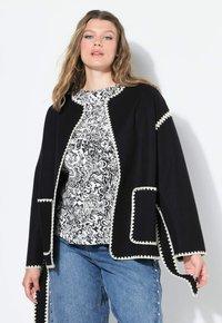 Femme portant un gilet noir avec des bordures blanches en zigzag par-dessus un haut à motifs noir et blanc et un jean bleu, se tenant contre un mur blanc.