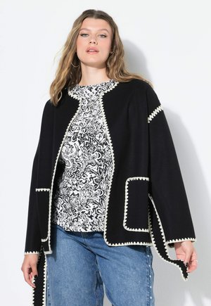 Frau in einem schwarzen Cardigan mit weißem Zickzackbesatz über einem schwarz-weißen Musteroberteil und blauen Jeans, stehend vor einer weißen Wand.