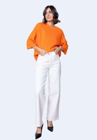 Maglione arancione a coste con maniche ampie, abbinato a jeans bianchi a vita alta e décolleté nere a punta. Sfondo chiaro e semplice.
