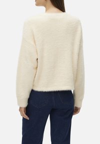 Un pull en peluche couleur crème avec une coupe décontractée et des manches longues, présentant des poignets et un ourlet côtelés. Porté avec un jean en denim noir.