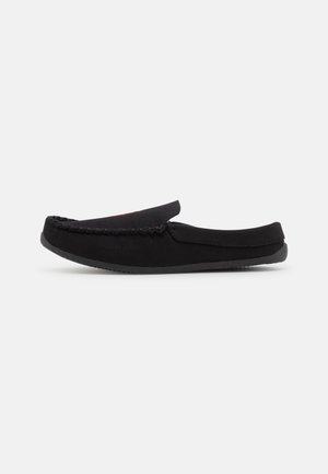 Zapato negro estilo mocasín sin cordones con detalles cosidos y suela de goma, mostrado de perfil sobre fondo liso.