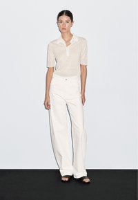 Beige ribbet strik polo skjorte med korte ærmer og krave, parret med vide hvide bukser og sorte slidesandaler på et sort gulv.