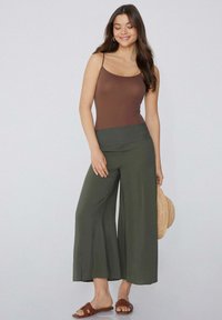 Hnědý střihový top na ramínka spárovaný s olivově zelenými širokými kalhotami se smockovaným pasem. Model nosí hnědé sandály a drží slamáky.