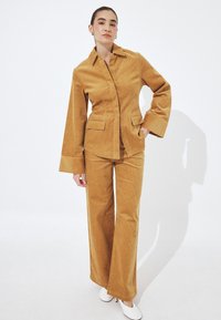 Woman standing in tan corduroy jacket and matching wide-leg pants with white heels against a plain white background.
