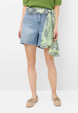 Kvinde iført lyseblå cowboyshorts med et grønt mønstret tørklæde som bælte, grøn striktop og beige mokkasinsko, stående mod en hvid baggrund.