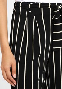 Zwart-witte gestreepte high-waisted broek met een strikceintuur, voorzien van plooien en een textuurstof. Hand zichtbaar ernaast.