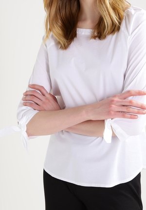 Blouse blanche semi-ajustée à manches trois-quarts, avec détail noué aux poignets et ourlet arrondi. Texture lisse et tissu léger.