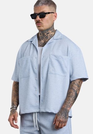 Mann mit Tätowierungen am Hals und an den Armen, der schwarze Sonnenbrillen, ein hellblaues kurzärmeliges Hemd mit Knöpfen, passende Shorts und ein weißes Tanktop trägt.