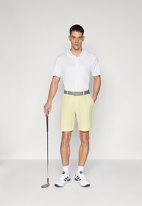 Gai dzelteni golfa šorti, balta polo krekls, pelēks josta, tur rokā golfa nūja. Balti apavi ar ziliem akcentiem un balti zeķes. Gluda auduma.