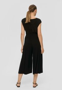 Zwarte jumpsuit met een geplooide stijl op de schouders, een wijde pijpen silhouet en een gladde stof, gestyled met platte zwarte sandalen.