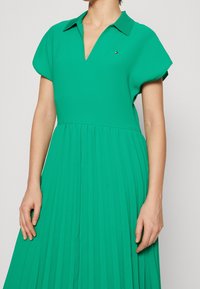 Vihreä poimutettu mekko, jossa on kaulus, V-pääntie ja lyhyet hihat. Materiaali näyttää sileältä, erittäin huomaamattomalla logoyksityiskohdalla rinnan lähellä.