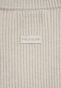 Elfenbeinfarbener, gerippter Strickstoff mit einem kleinen rechteckigen "POLO CLUB"-Etikett, das mit passendem Garn eingenäht ist.