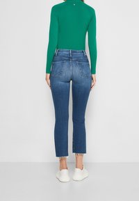 Högmidjade blå denimjeans med rak benpassform och fransad fåll, i kombination med en grön långärmad tröja och vita sneakers.