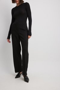Schwarzes Langarmoberteil mit einer engen Rippstruktur, kombiniert mit lockeren schwarzen Hosen. Das Modell trägt spitze schwarze Pumps.