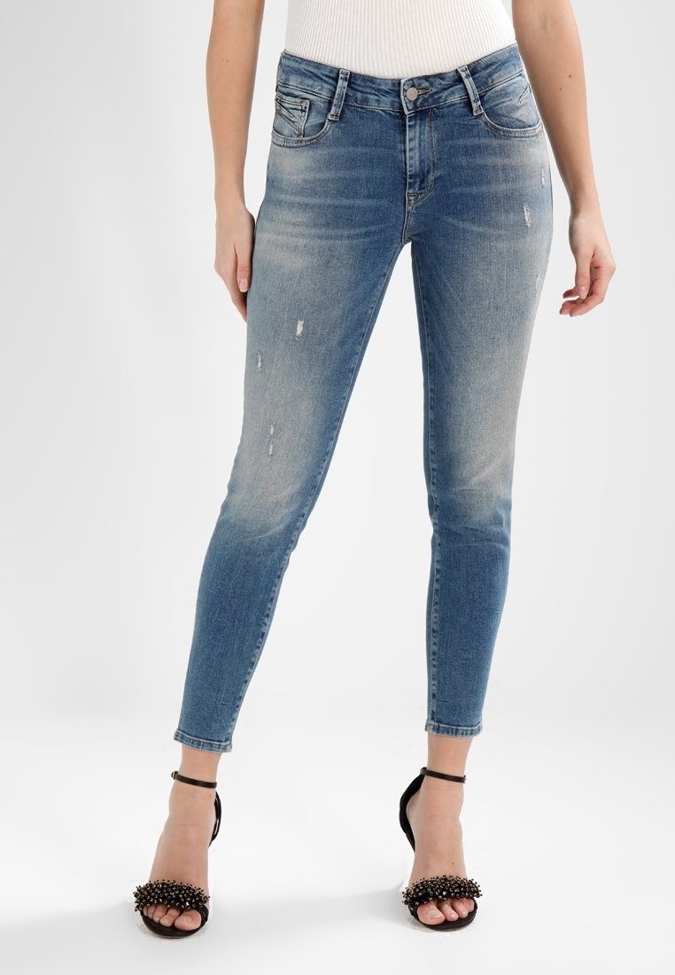 Femme portant un jean skinny bleu délavé et des sandales à talons hauts noirs avec des brides à la cheville et des clous décoratifs.