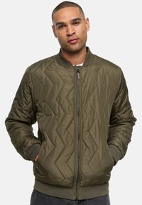 Chaqueta bomber acolchada en verde oliva con cremallera frontal, puños y dobladillo acanalados; presenta un patrón en Chevron y un diseño clásico.