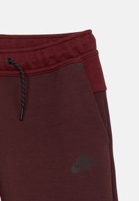 Tamsiai raudoni Nike treningo kelnės su tekstūruotu juosmeniu, turinčiu juodą raištelį ir logotipą. Audinys atrodo minkštas ir patogus.