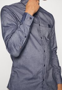 Lightweight, dark blue long-sleeve shirt with a button-down collar, featuring a chest pocket and subtle texture. Blue buttons and an orange accent.