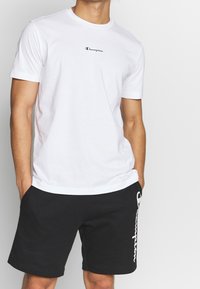 Hvid bomulds t-shirt med et lille sort Champion logo på brystet, parret med sorte shorts med et hvidt Champion logo på siden.
