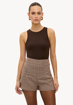 Vrouw met naar achteren gekamd haar draagt gouden oorringen, een nauwsluitende mouwloze bruine top, bruine geruite shorts met hoge taille en doorzichtige donkere panty's.