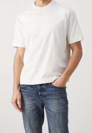 White cotton t-shirt featuring a raised logo design on the chest, short sleeves, and a high collar, paired with blue distressed jeans.
