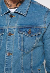 Modrá džínová bunda s dvěma kapsami na hrudi, měděnými knoflíky a žlutým šitím, s klasickým límečkem a lehce vybledlým vzhledem.