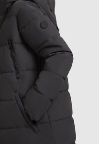 Black insulated puffer jacket featuring a zippered chest pocket, side pockets, and a round logo patch on the left sleeve. Smooth texture.