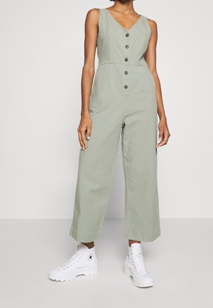 Person iført ærmeløs salviegrøn jumpsuit med knapper foran, vide ben og hvide højtop-sneakers, stående på gråt gulv.