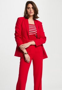 Rode suit met gestructureerde blazer en wijde broek. Daaronder een gestreept rood-wit topje. Glad materiaal, minimalistisch ontwerp, gouden manchetarmband.