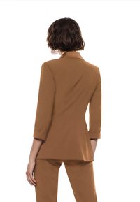 Blazer marrone su misura con una silhouette aderente, maniche lunghe e polsini arrotolati. Tessuto liscio e dettaglio del colletto affilato.