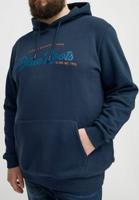 Sötétkék kapucnis pulóver puha anyagból, elől zsebbel és húzózsinórral. Az elülső részen a "Blue Roots" felirat látható világoskék és narancssárga színben.