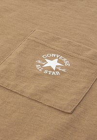 Rudas audinys su kvadratiniu kišenėmis. Baltas „CONVERSE ALL STAR“ logotipas apskritime su žvaigžde centre.