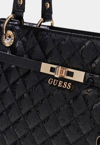 Borsa a tracolla nera trapuntata con finitura lucida, dotata di una fibbia in metallo dorato e logo "GUESS" su una cintura orizzontale. Doppi manici per la spalla.