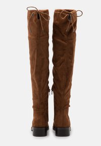 Bottes en daim brun au-dessus du genou avec des liens ajustables en haut. Elles présentent un design élégant et un petit talon carré pour plus de stabilité.