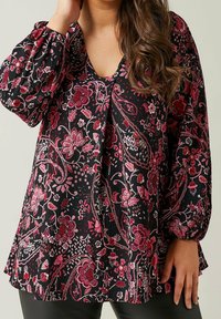 Blouse noire à motifs floraux avec un col en V, des manches longues bouffantes, et un motif texturé dans des tons de rouge et blanc. L'ourlet est légèrement évasé.