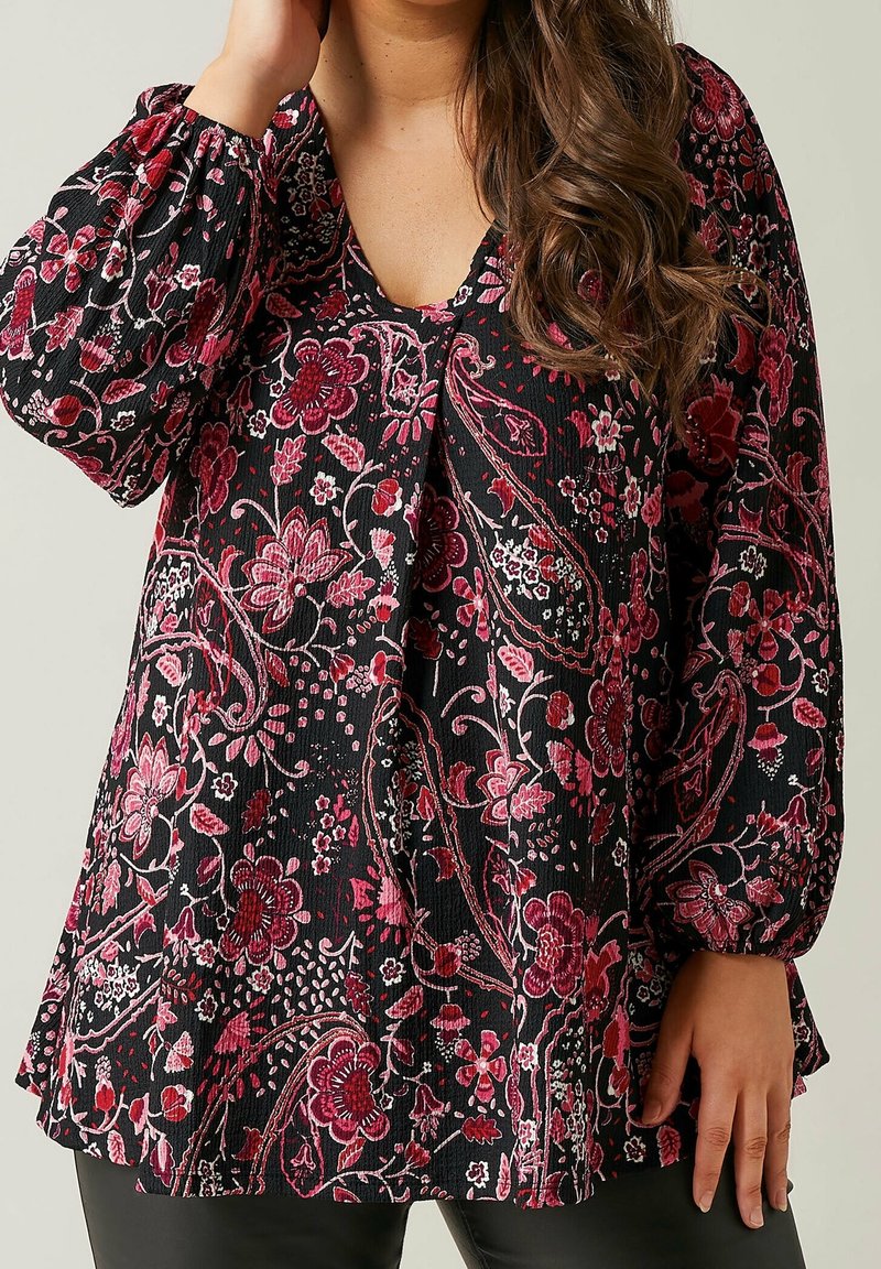 Blouse noire à motifs floraux avec un col en V, des manches longues bouffantes, et un motif texturé dans des tons de rouge et blanc. L'ourlet est légèrement évasé.
