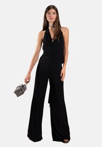Haut noir à col halter avec un design drapé, associé à un pantalon noir à jambes larges. Les accessoires comprennent un petit sac à cordon gris et un col métallique.