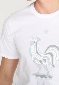 Valkoinen t-paita, jossa on tyylitelty kukko -grafiikka harmaana ja sinisenä, aaltoilevilla viivoilla ja tähti päällä, rintakappaleella.