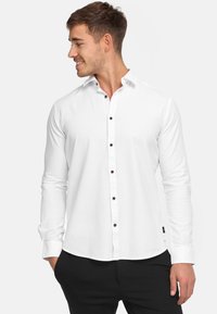 Weißes Langarmhemd mit Button-Down-Kragen, ausgestattet mit braunen Knöpfen, einer glatten Textur und einem abgerundeten Saum, kombiniert mit schwarzen Hosen.