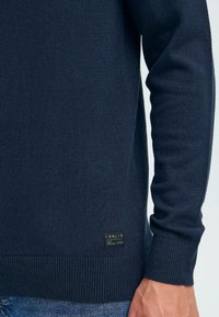 Pulover tricotat de culoare bleu navy, cu manșete și tiv ribalte, având o etichetă cu logo mic pe partea stângă jos. Textură moale și design ajustat.